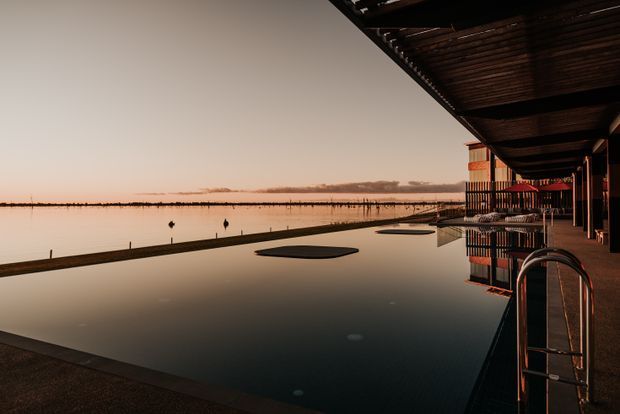 A tranquil waterfront exterior at sunset, showing a long reflective pool beside a covered building veranda or promenade. Dark timber roof framing and posts line the right side, with pool ladders and a distant calm body of water beyond.