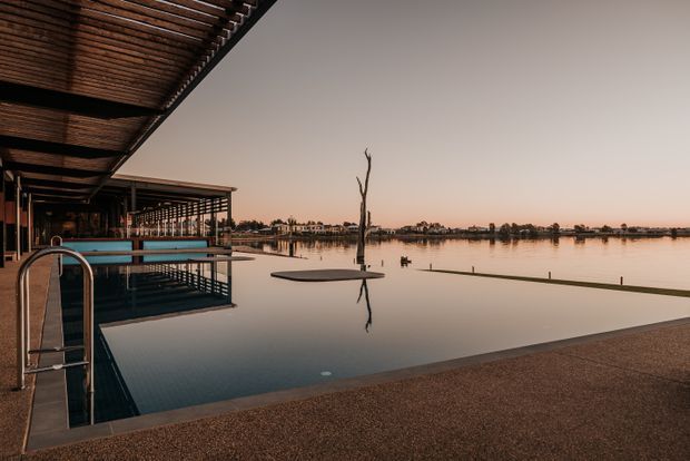 A tranquil waterfront pool terrace at dusk, with a dark timber-clad covered structure and slatted roof projecting over the pool. The infinity pool reflects the calm lake, while a sculptural dead tree rises from the water and low buildings line the distant shore.