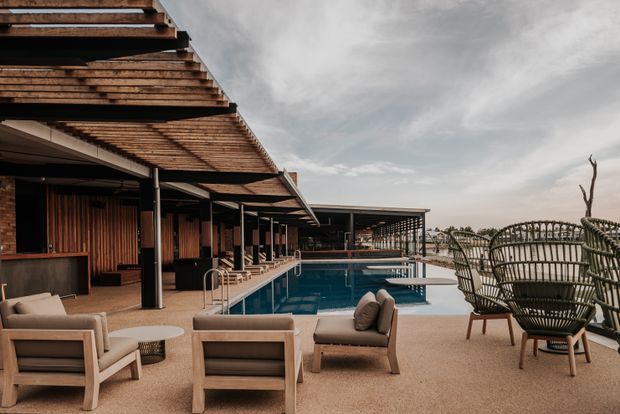 A sophisticated outdoor pool terrace beside a low-rise pavilion, with timber pergola shading, dark structural posts, warm timber wall cladding, outdoor lounge seating, and a long rectangular swimming pool. The terrace overlooks a bright waterfront or coastal setting, creating a resort-like atmosphere.