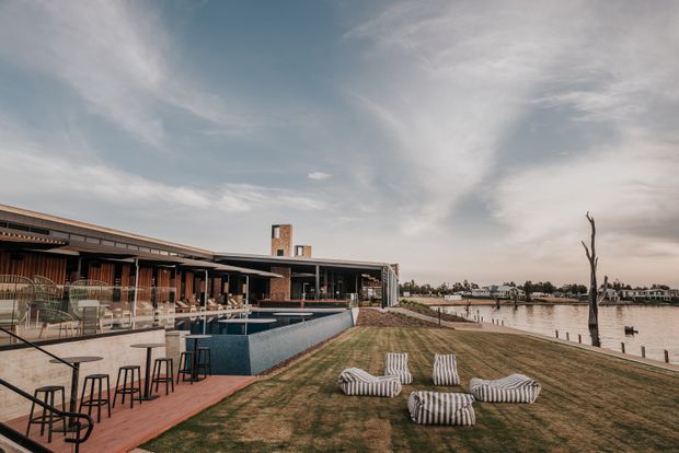 A contemporary waterfront property with a long, low-slung building opening onto a pool terrace and landscaped lawn. The outdoor area includes a bar-height dining setting, lounge chairs, and a swimming pool beside a calm body of water. The architecture features extensive glazing, dark horizontal rooflines, timber-toned walls, and a prominent brick chimney.