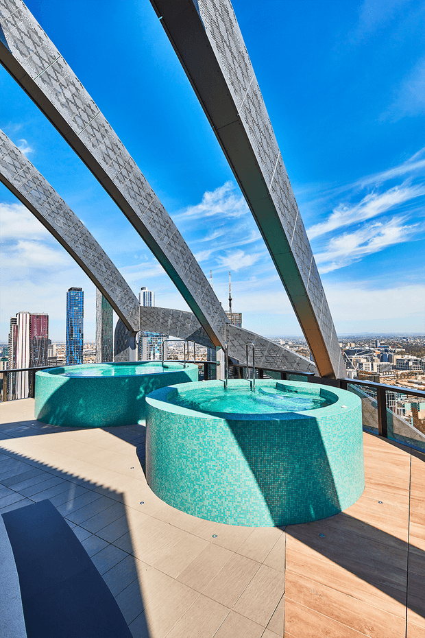 A rooftop terrace high above a dense city skyline, featuring two round turquoise mosaic-tiled spa pools or plunge pools on a large tiled and timber-look deck. Dramatic angled structural steel beams frame the view, with glass balustrades, bright blue sky, and strong sunlight creating bold shadows.
