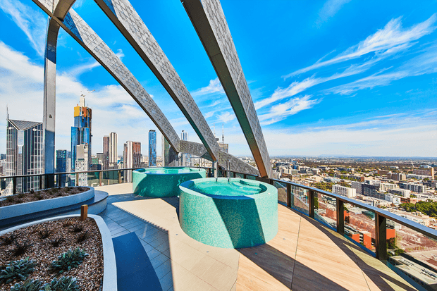 A high-rise rooftop terrace overlooking a dense city skyline, featuring two turquoise tiled circular spa or plunge-pool structures, curved built-in planters with succulents, timber decking, dark paving, glass balustrades, and dramatic arched structural beams overhead. The scene is bright and contemporary with expansive urban views.