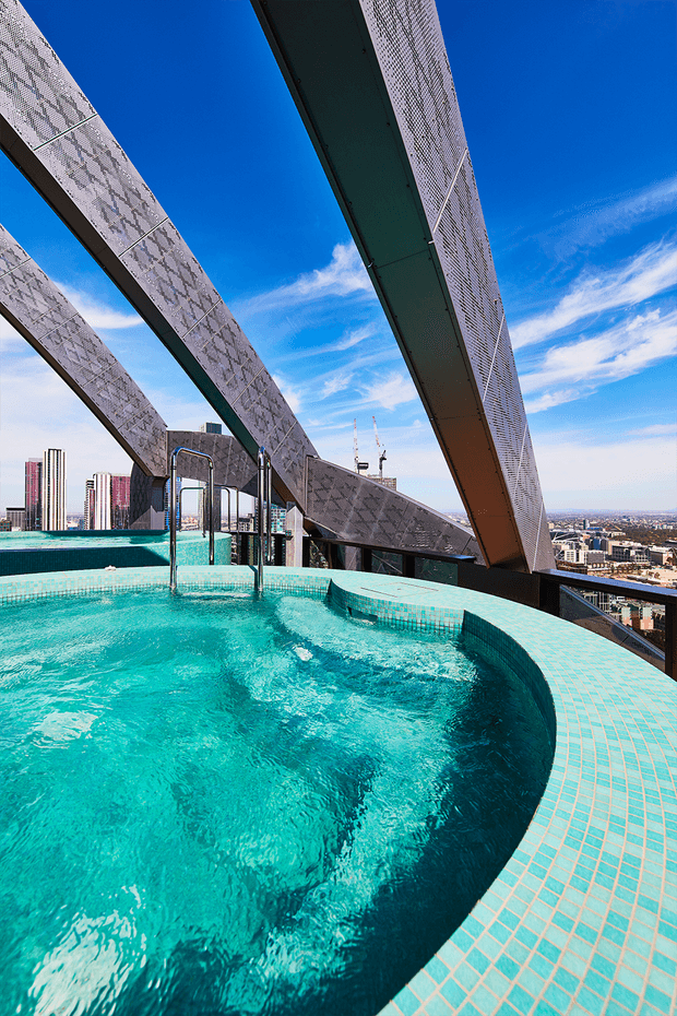 A rooftop pool or spa with vivid turquoise water and a curved, small-format turquoise tiled edge, set high above a dense urban cityscape. Dramatic angled steel architectural structures frame the pool against a bright blue sky.