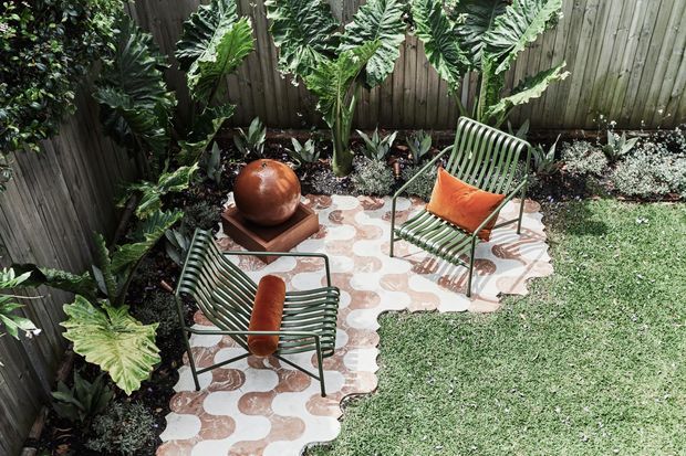 A landscaped courtyard garden with a patterned tiled path or patio, two green metal outdoor lounge chairs, and a small central pedestal table with a rounded terracotta-toned ceramic vessel. Dense tropical foliage borders the space, with a timber privacy fence enclosing the garden and lawn.