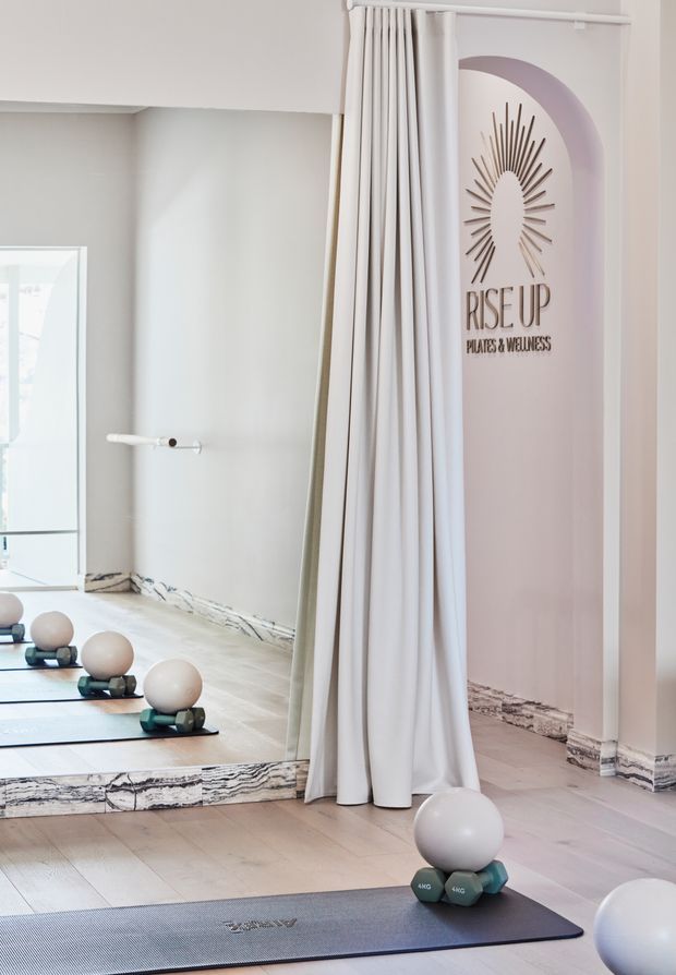 Bright, softly styled Pilates and wellness studio with pale wood flooring, light neutral walls, full-height mirrors, white curtains, exercise mats, small weights, and Pilates balls. A blush-toned arched wall feature carries the “Rise Up Pilates & Wellness” branding. The space feels clean, airy, and calm.