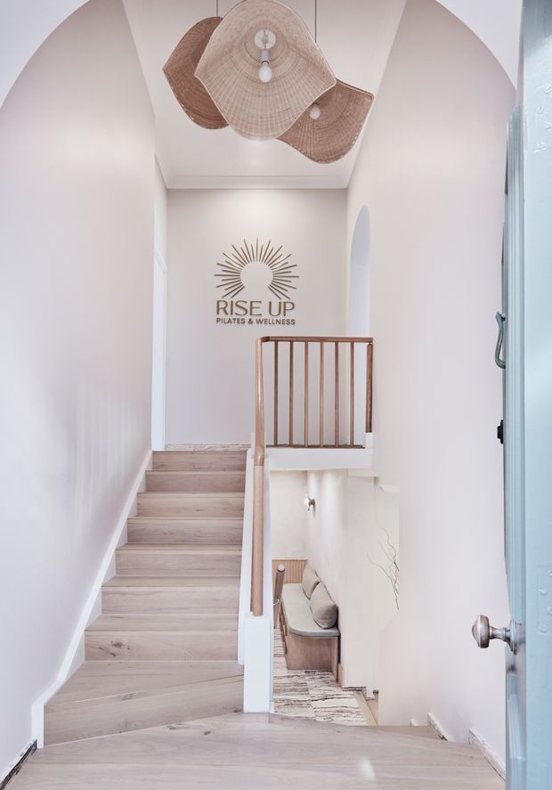 Bright, refined stairway and landing with pale timber stair treads, white walls, arched openings, and a warm timber balustrade. A sculptural woven pendant light hangs overhead, while a Pilates and wellness sign is mounted on the upper wall. The lower level includes a small seating nook visible beneath the stairs.