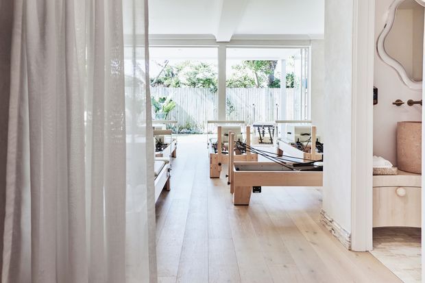 Bright, airy Pilates or reformer studio viewed through a wide opening, with multiple wooden reformer machines arranged on pale timber flooring. The room has large windows with sheer curtains, white walls and ceiling beams, and built-in cabinetry visible at the right edge.