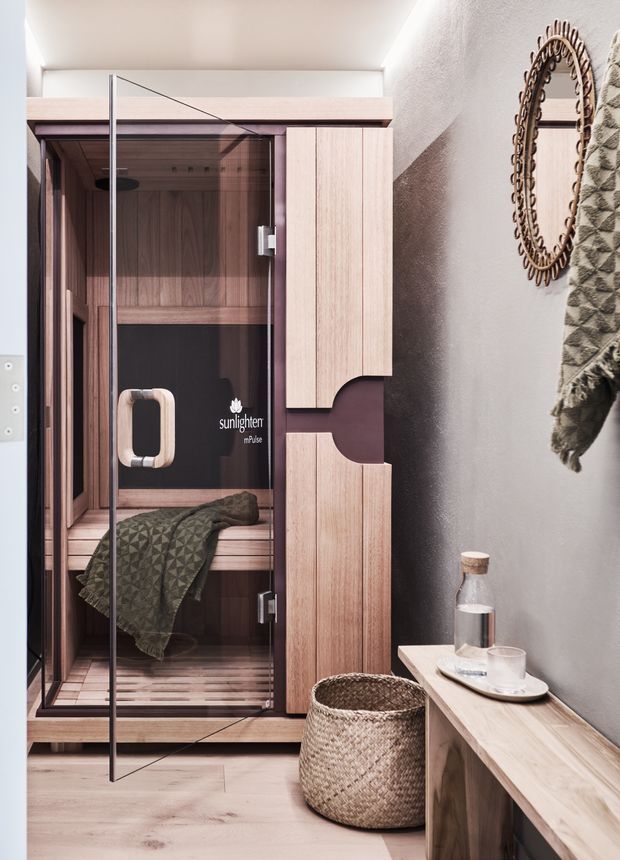 A compact home sauna with a clear glass door, warm timber interior and built-in timber cabinetry. A textured grey wall, decorative oval mirror, woven basket, bench console and folded patterned textile create a calm, spa-like setting.