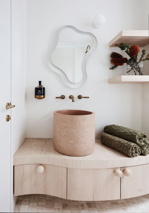 A softly styled bathroom vanity with a round pink-beige stone vessel basin, wall-mounted aged brass tapware, an irregular organic-shaped mirror, pale timber curved-front cabinetry, and open timber shelves displaying dried botanical arrangements. The room has light neutral walls, a curved stone benchtop, and patterned floor tiles.