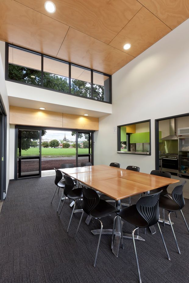 A bright double-height communal dining and meeting space with a large timber table surrounded by black chairs. Dark carpet covers the floor, while white walls contrast with a warm timber-panelled ceiling and matching timber joinery. Full-height black-framed glazing and an upper-level window band overlook a green open area. A compact kitchen servery is visible through an internal opening on the right.