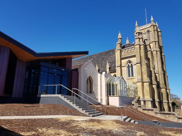 Exterior view of a contemporary dark-clad building addition beside a historic stone Gothic church. The modern structure has a sharply angled roof, extensive dark glazing, timber-toned soffits, and an accessible stair and ramp approach. The church features pointed-arch windows, buttresses, crenellated detailing, and multiple spires. Clear blue sky and landscaped grounds surround the buildings.