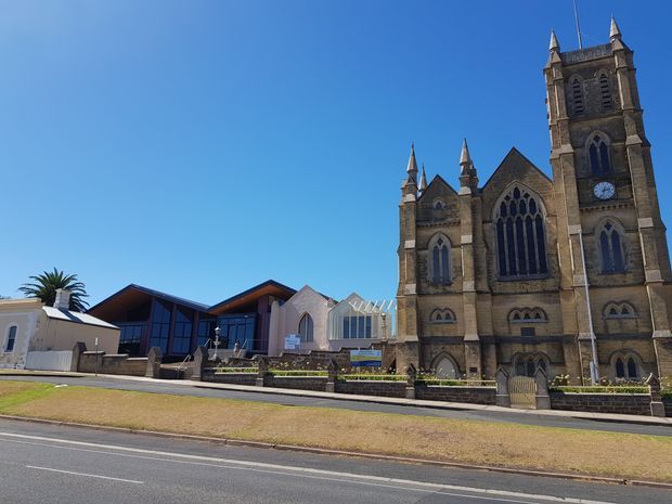 Street-level exterior view of a large Gothic Revival stone church with a tall twin-towered facade, pointed-arch windows, pinnacles, clock, and decorative masonry. A contemporary angular-roof building is visible beside it, with a paved frontage, low stone walls, lawn, road, and clear blue sky.