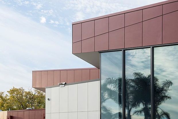 Exterior architectural detail of a contemporary, rectilinear building with projecting box forms clad in muted reddish-brown panels, white wall panels, and tall dark-framed glazing reflecting palm trees and sky. The composition emphasizes cantilevered volumes, crisp grid lines, and a flat-roof profile.