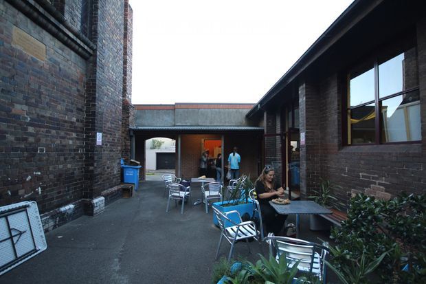 A narrow enclosed courtyard or laneway between dark brick buildings, furnished with outdoor café tables and metal chairs. Large windows and covered roof edges line the right side, while an arched passage and doorway connect to interior spaces at the rear. Potted plants soften the industrial masonry setting, with people seated outdoors.