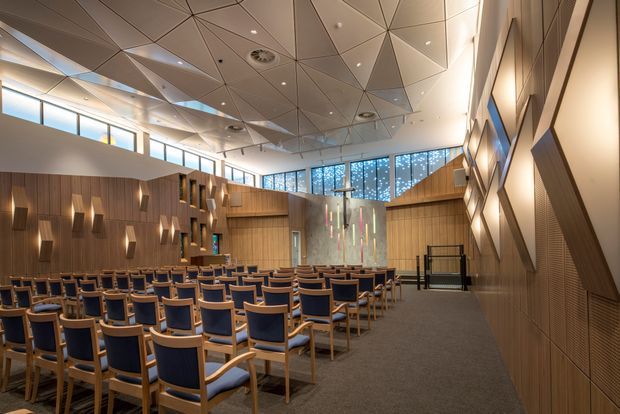 A large contemporary auditorium or conference hall with rows of blue upholstered chairs, extensive warm timber wall panelling, a raised presentation area, and decorative vertical light elements. The ceiling features a dramatic geometric faceted design with integrated recessed lighting, while clerestory glazing and patterned upper windows bring in daylight. The space has a refined, institutional or commercial character.