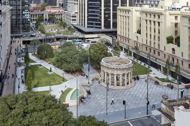 Elevated view of a formal urban civic plaza surrounded by dense multi-storey commercial and institutional buildings. The landscaped square includes lawns, mature trees, paved pedestrian paths, a circular classical colonnade, a shallow ornamental water feature, and surrounding streets with pedestrians. The architecture combines modern glass-and-steel office towers with a prominent neoclassical stone-clad building.
