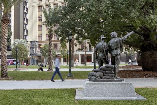 Urban landscaped public courtyard or plaza with mature trees, lawn, paved walkways, decorative street lamps, and a prominent bronze statue group on a raised stone pedestal. Multi-storey apartment or commercial buildings with detailed façades and balconies form the background, with pedestrians moving through the space.