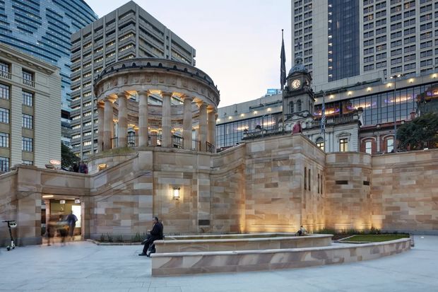 An urban civic plaza framed by monumental sandstone and stone-clad architecture. The scene features a semicircular classical colonnade with Corinthian-style columns, broad steps, illuminated retaining walls, a formal raised planter, and a historic clock tower integrated among surrounding high-rise office buildings. The dusk setting and architectural lighting create a refined, dramatic atmosphere.