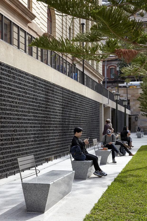 A landscaped urban courtyard or public forecourt beside a substantial civic or commercial building. A long dark textured wall carries an integrated text-based artwork, while a pale concrete path is furnished with angular granite benches and simple metal chairs. Mature cycad-like planting, lawn, and black lantern-style wall lights soften the formal masonry architecture. Several people are seated and walking through the space.