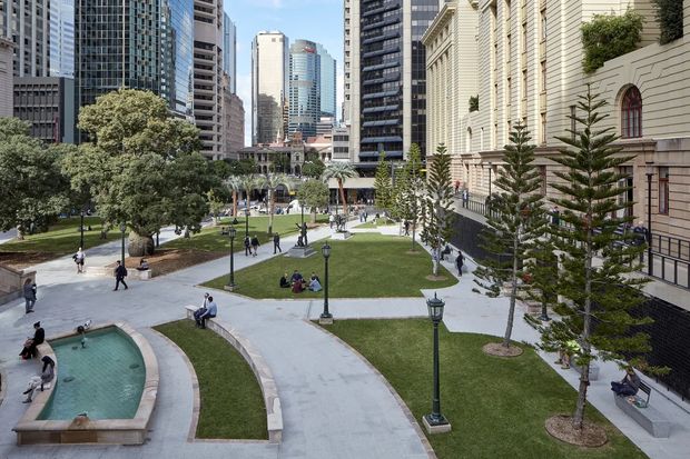 A landscaped urban civic courtyard or plaza viewed from an elevated position, with curved pale paving paths, clipped lawns, mature trees, ornamental conifers, traditional lantern-style light poles, and a shallow curved water feature in the foreground. A large ornate historic-looking stone building with arched windows occupies the right side, while dense modern high-rise buildings form the surrounding city backdrop.