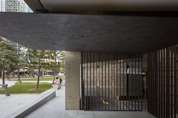 A covered urban entrance or passage beneath a broad concrete canopy, with a tall vertical metal security screen enclosing a glazed doorway. Concrete columns, soffit, and paved flooring create a monolithic institutional or commercial character. A landscaped public plaza and high-rise buildings are visible beyond the opening.