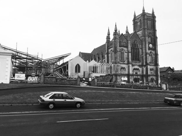 Black-and-white street-level view of a large historic Gothic Revival church with twin pointed towers, ornate stone masonry, arched tracery windows, pinnacles, and a steeply pitched roof. A smaller adjoining structure and a modern construction site are visible beside it, with a roadway and parked cars in the foreground.