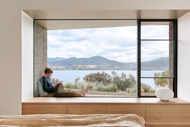 A serene bedroom interior with a large black-framed picture window overlooking a lake and mountainous landscape. A person sits in a deep timber-framed window seat, while a broad built-in timber storage platform runs beneath the glazing and the foreground shows part of a bed. The room has pale plaster walls, exposed light brick reveals, warm timber joinery, and a calm minimalist aesthetic.