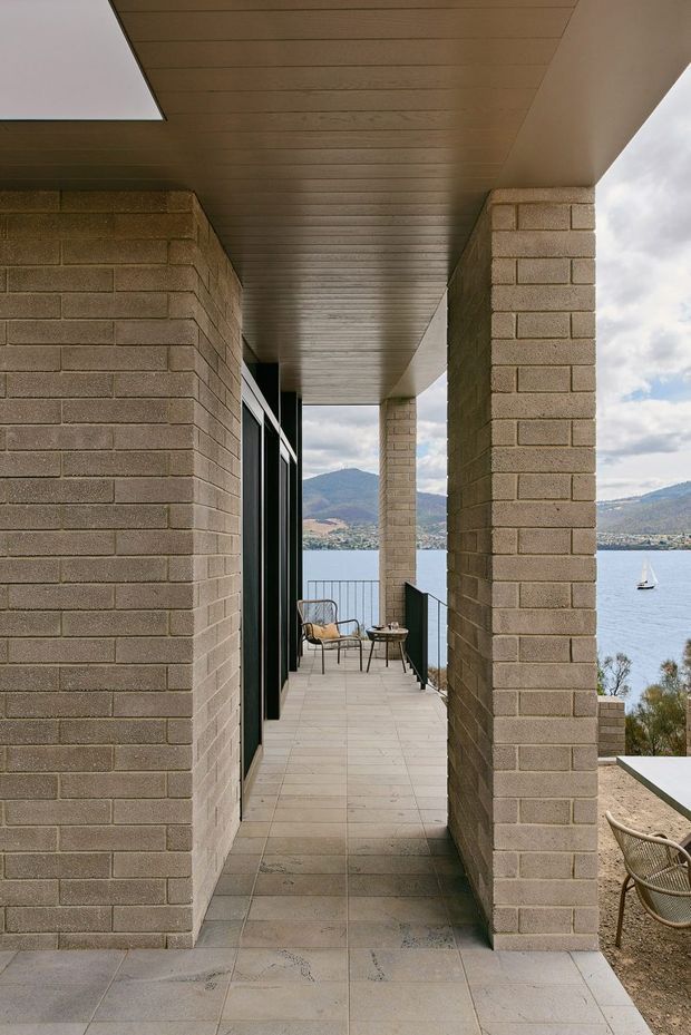 A covered exterior balcony or terrace corridor framed by substantial tan brick piers, with a horizontal timber-lined soffit and large dark-framed glazing. Pale tiled paving leads to a small outdoor seating area overlooking a lake, sailboat, and mountainous shoreline. The image emphasises the sheltered outdoor circulation space, strong masonry structure, and waterfront outlook.