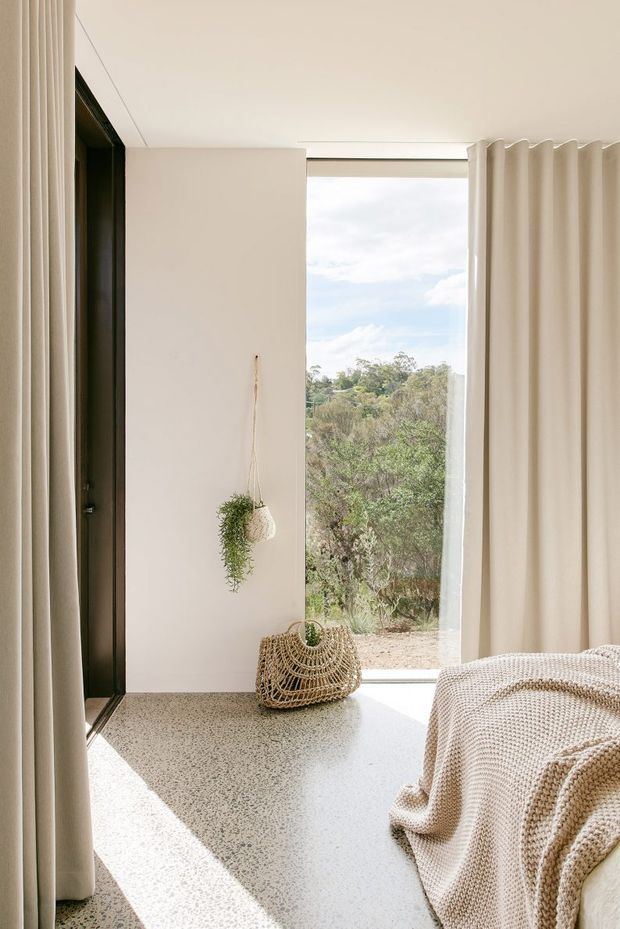 A bright, serene bedroom corner with floor-to-ceiling glazing opening onto a natural landscape. Soft cream curtains frame the view, while a textured knit throw covers the edge of the bed. The room has pale walls, a speckled light terrazzo-style floor, dark window framing, and restrained woven and botanical accents.