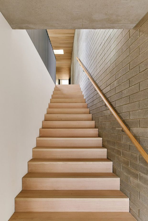 A narrow modern stairway viewed from the bottom, with broad light timber treads and risers ascending toward a small upper landing. One side has a textured exposed concrete-block wall with a simple timber handrail, while the opposite side is smooth white plaster. The ceiling is raw concrete, with a warm timber-lined section and a dark vertical balustrade visible near the top. Natural and overhead light create a restrained, material-focused interior.