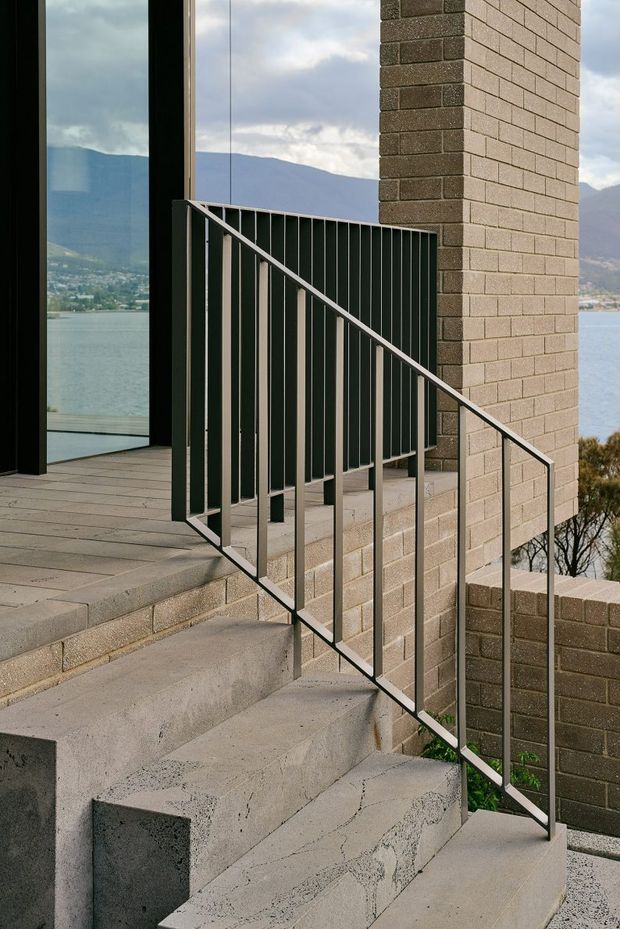A close architectural view of an exterior concrete stairway and landing beside a tall textured brick wall. Slim dark metal balustrades frame the stairs, with large dark-framed glazing at the upper landing. A lake and mountainous landscape are visible beyond the building, reinforcing the elevated outdoor setting.