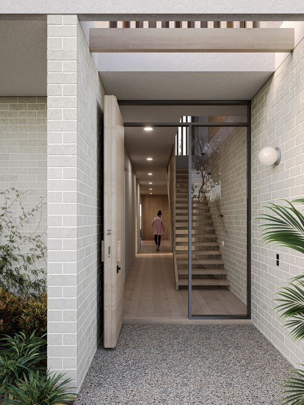 A contemporary residential entry sequence viewed from a small landscaped courtyard. The foreground has a speckled terrazzo-like paved floor, pale grey brick walls, and tropical planting. A glazed dark-framed door opens to a long timber-floored hallway, with a timber staircase, recessed ceiling lights, and a glazed internal partition. Exposed beams and a patterned upper screen add architectural detail.