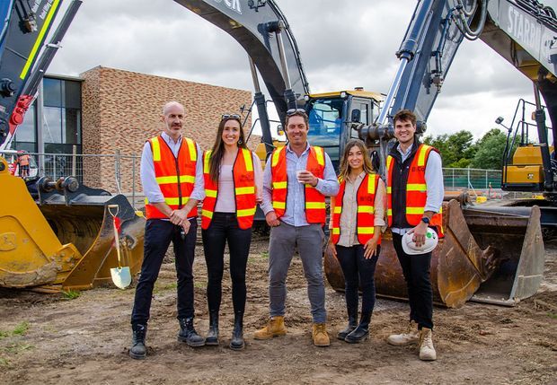 A group of five construction professionals wearing high-visibility safety vests pose in front of large excavators and earthmoving equipment at an active construction site. A brick building and temporary site fencing are visible in the background under an overcast sky.
