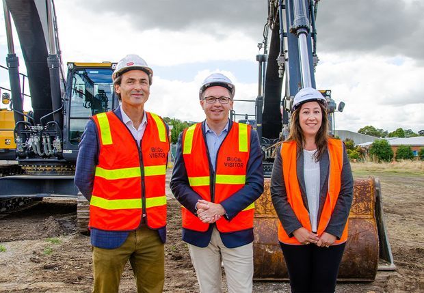 Three construction site visitors wearing hard hats and high-visibility safety vests stand in front of heavy excavation machinery and a large bucket. The image is an outdoor construction-site portrait with industrial equipment, disturbed ground, and low-rise buildings in the background.