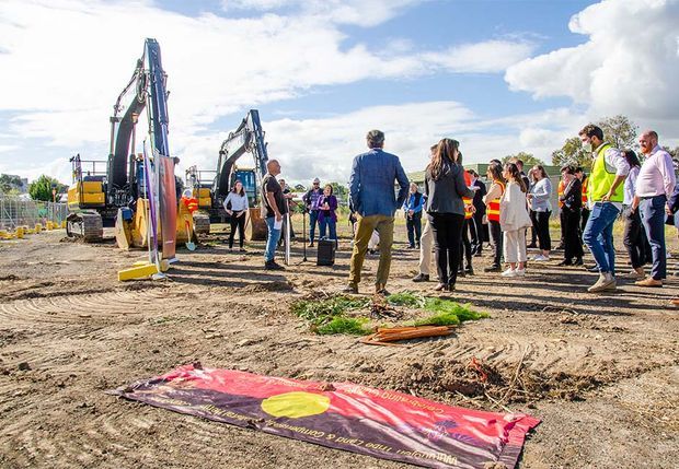 An outdoor construction site with excavators, safety fencing, and a group of people gathered for what appears to be a ground-breaking or project launch event. A large ceremonial banner lies on the dirt in the foreground under a bright, partly cloudy sky.