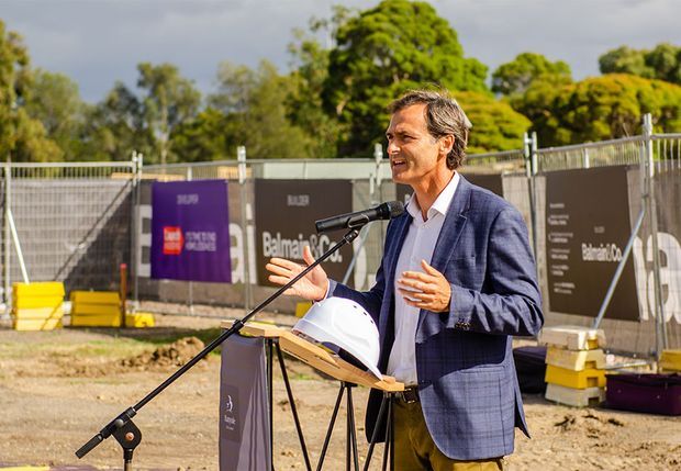 An outdoor construction-site event showing a suited man speaking at a microphone behind a lectern, with a white hard hat and construction hoarding in the background. Trees and site materials surround the temporary presentation area.