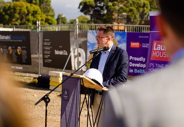 Outdoor event photograph showing a man in a suit speaking at a podium with a microphone, surrounded by construction-site fencing and development signage. Attendees are partially visible in the foreground, with trees and an active or upcoming development site in the background.