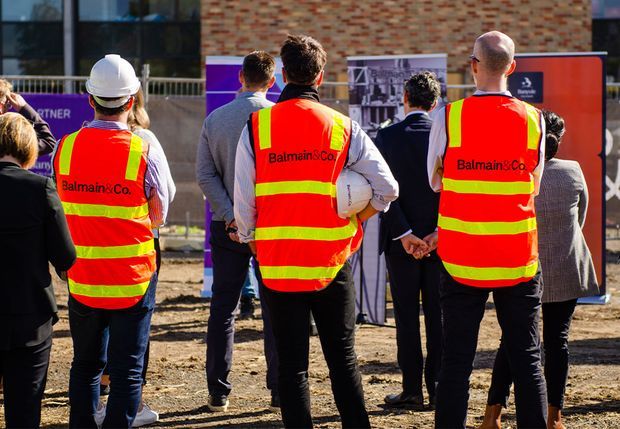 Outdoor construction-site event with several people viewed from behind, including workers wearing high-visibility red and yellow safety vests and a white hard hat. Promotional signage, a brick building facade, temporary fencing, and exposed ground are visible in the background.