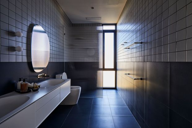 A contemporary bathroom with a long floating double vanity, integrated basins, wall-mounted faucets, and a rounded illuminated mirror. The room is finished in dark grey tiled walls and floor, with a white toilet and a narrow full-height glazed door or window at the far end. Linear wall-mounted towel rails and restrained lighting create a sleek, moody atmosphere.