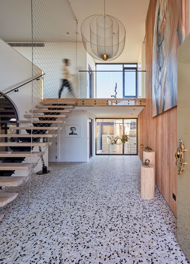 A bright, double-height residential entry hall with a dramatic floating staircase, cable-net balustrade, timber mezzanine bridge, and oversized sculptural pendant light. The space features a terrazzo floor, white walls, timber wall panelling, large windows, and a glazed rear courtyard connection. A large abstract artwork and decorative objects add visual interest.