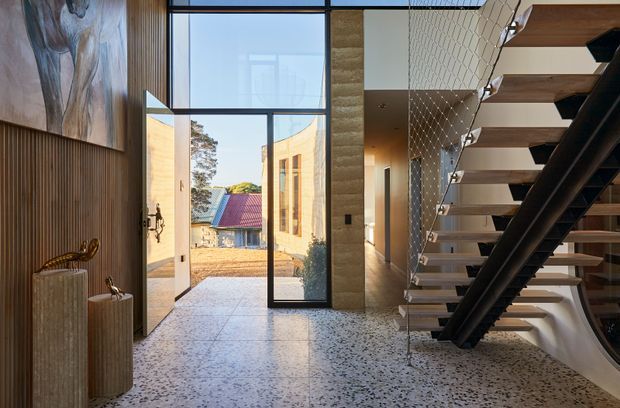 A refined contemporary entryway with a terrazzo floor, tall floor-to-ceiling glazing and a pivoting front door opening to an exterior courtyard. Vertical timber wall panelling, sculptural pedestal basins, a stone-textured wall and a striking open-riser staircase with dark structural elements define the space. An upper-level void or mezzanine edge with wire balustrading is visible above the stair.