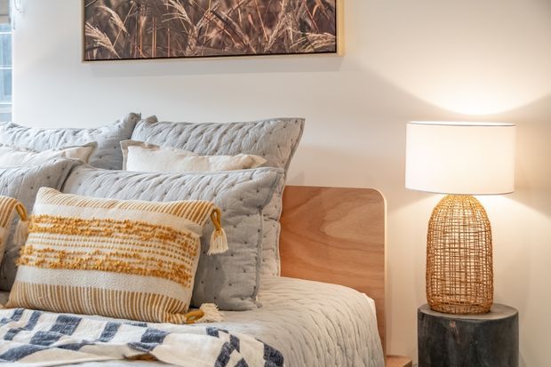 A warmly styled bedroom corner featuring a layered bed with quilted light-grey bedding, textured cream and mustard cushions, and a blue-and-white throw. A rounded timber bedside table sits beside a woven basket-style table lamp on a dark cylindrical pedestal. Neutral walls, soft lighting, and botanical artwork create a calm, cozy atmosphere.