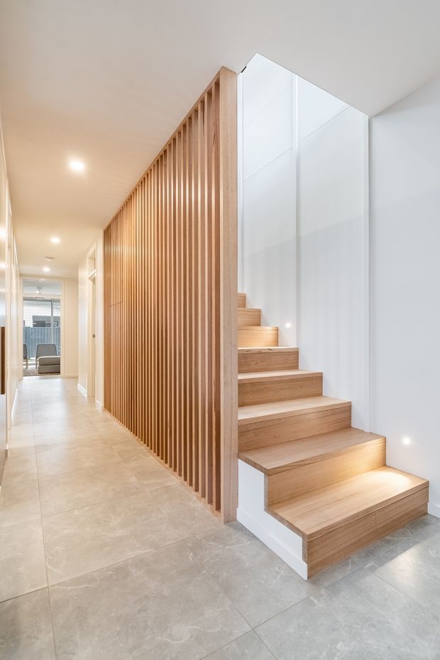 Bright contemporary residential hallway featuring a timber staircase with open risers and integrated under-step lighting. A full-height vertical timber batten screen separates the stair from the corridor, while large light-grey stone-look floor tiles, white walls, and recessed ceiling lights create a clean, refined interior.