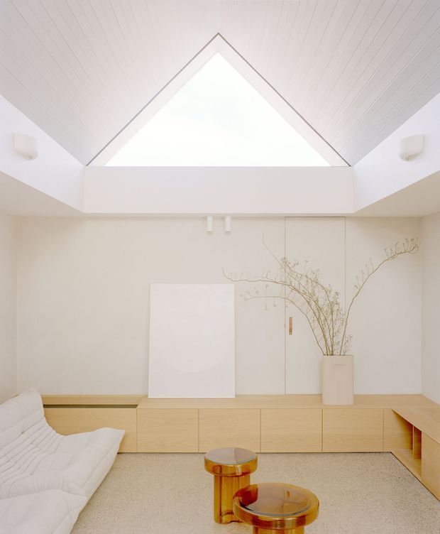 A bright minimalist living room with a warm pale timber built-in storage wall and low media console, a white sculptural lounge chair, and amber glass coffee tables. The room features a dramatic pitched ceiling lined with narrow white timber boards and a large triangular clerestory or skylight-like opening, creating an airy, gallery-like atmosphere. Neutral plaster walls, concealed cabinetry, and sparse sculptural decor reinforce the refined minimalist design.