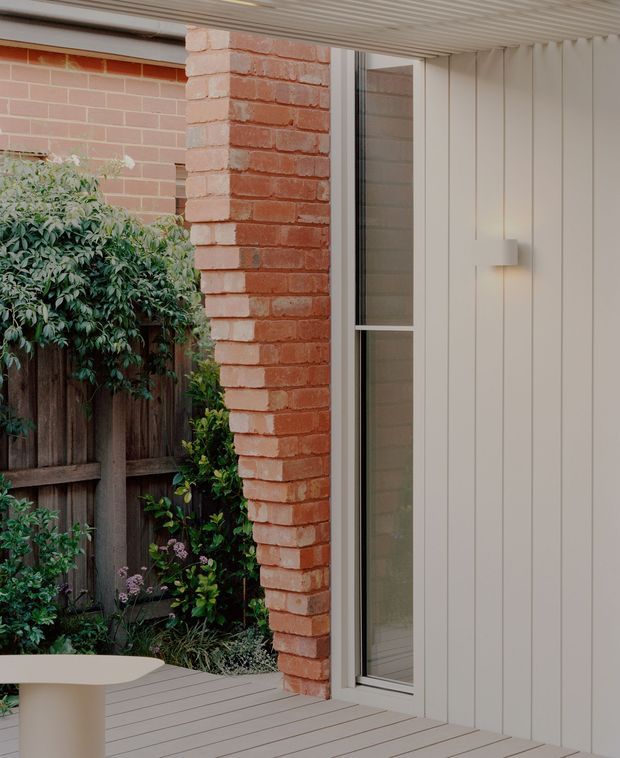 Close-up architectural view of a covered exterior deck or patio beside a pale vertical-panelled wall. A red brick structural pier frames a tall narrow glazed door or window, with a timber-framed garden fence and dense planting visible beyond. The deck has light grey timber boards, and a small wall-mounted light is installed on the panelled facade.