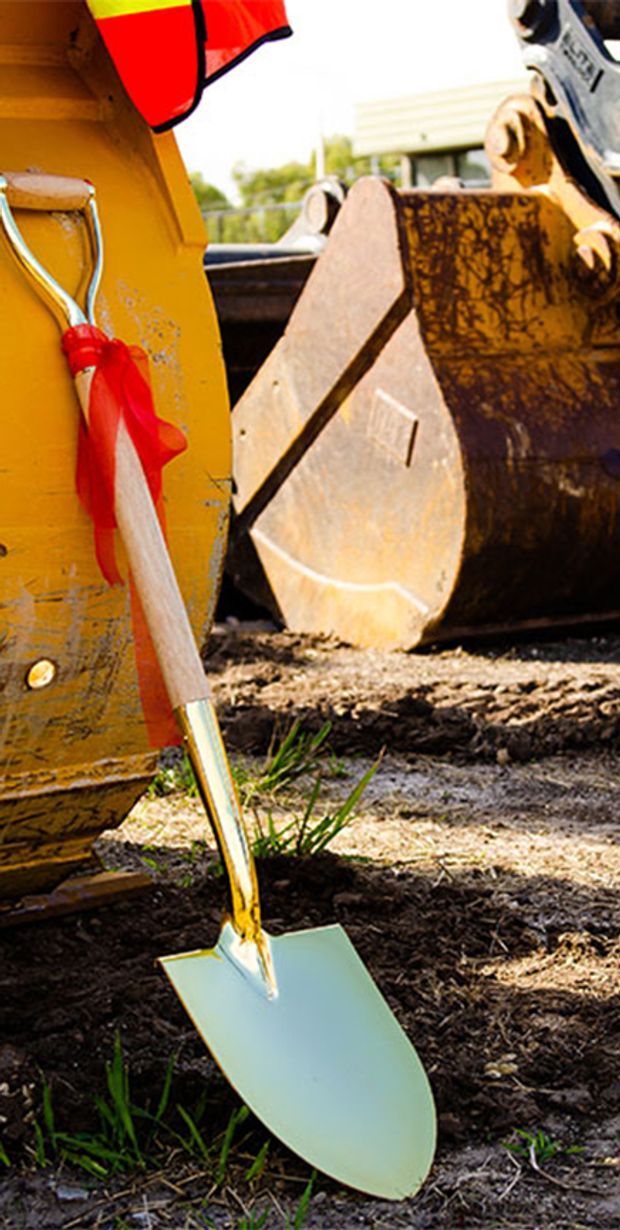 Close-up outdoor construction scene showing a shovel planted in disturbed soil beside the rusted bucket and yellow body of an excavator. A high-visibility red and yellow garment is partially visible, with construction equipment and a blurred site background.