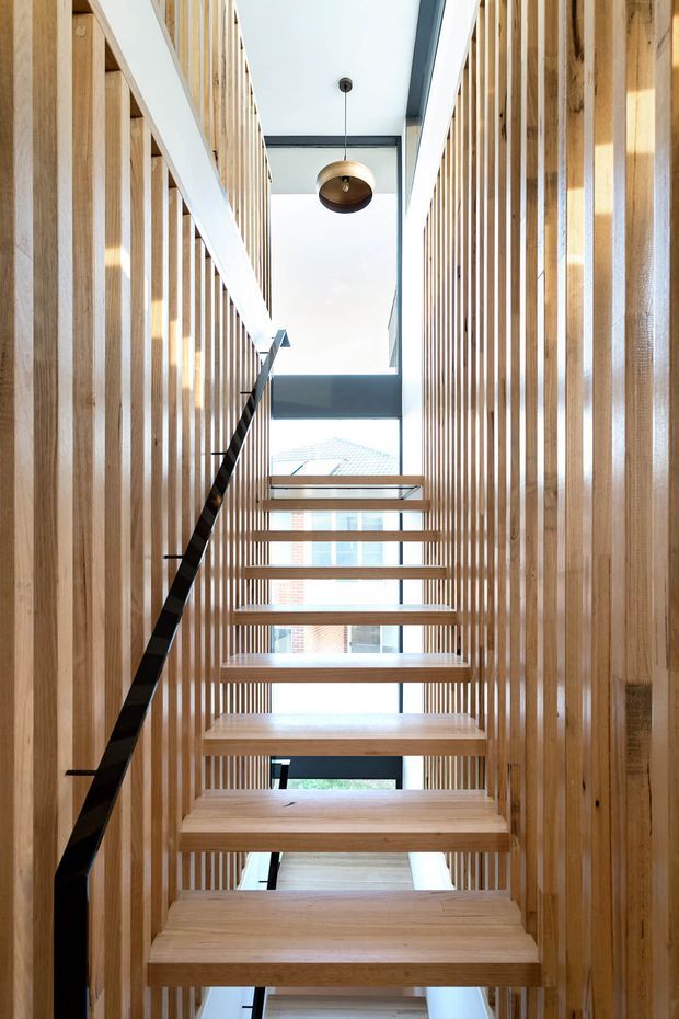 Interior view looking up a narrow, light-filled stairway with open-riser timber stairs, black metal handrail, extensive vertical timber slat screening on both sides, and a high glazed opening at the landing. A simple suspended pendant light hangs beneath a white ceiling with dark structural beams.