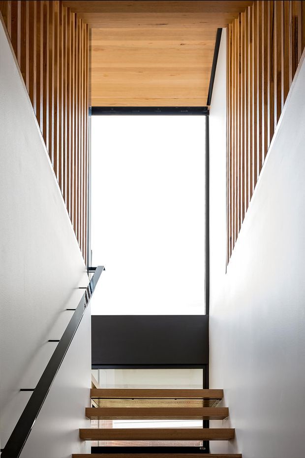 A bright contemporary stairway viewed from below, with floating timber treads, slim black metal balustrade, white walls, and a dramatic vertical glazed opening. Vertical timber battens line both sides beneath a warm timber ceiling, creating a strong geometric and material-focused composition.