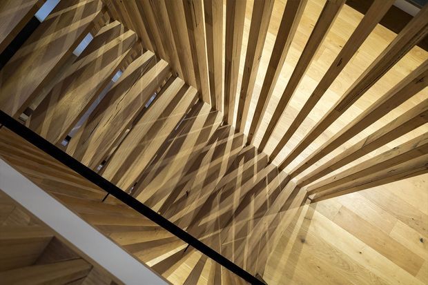Dramatic upward view of a timber stairwell or stair void, with numerous exposed solid-wood stair treads and structural beams forming a radial, sculptural pattern around the opening. A dark metal or black balustrade crosses the foreground, while skylight-like openings bring in natural light and cast strong geometric shadows across the timber surfaces.