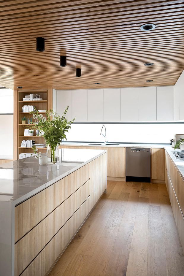 Bright contemporary kitchen with a large central island, pale timber cabinetry, white upper cabinets, and a light stone benchtop. The room features extensive built-in joinery, integrated appliances, open shelving, a timber slatted ceiling with recessed and pendant lighting, and wide timber flooring. A large window or glazed opening brings in abundant natural light, while greenery and books add a natural, lived-in touch.